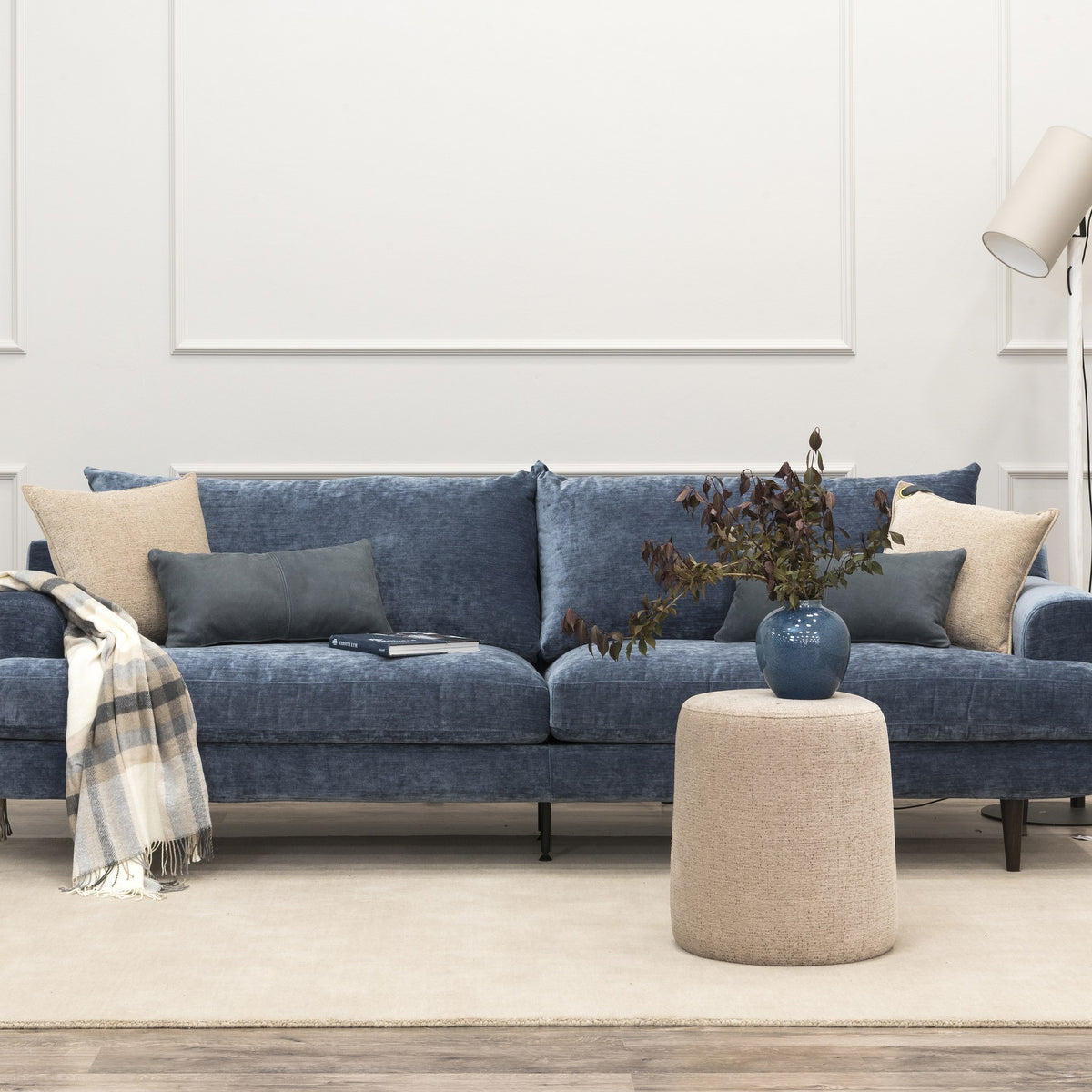 Leon sofa i denimblå chenille i lyst rom med panelvegg, beige puff og rutete pledd