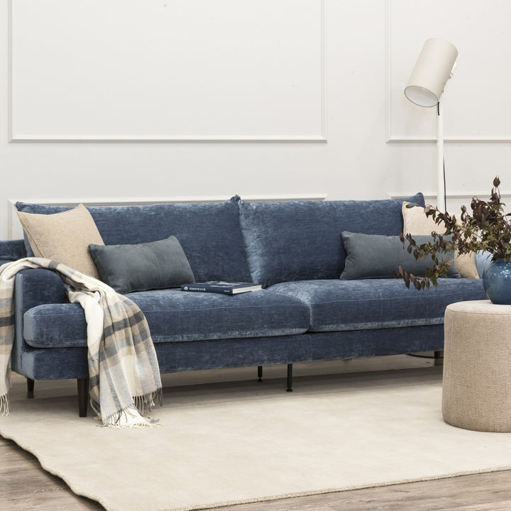 Leon sofa i blå chenille i lys stue med pledd, puter, gulvlampe og vase med grener.