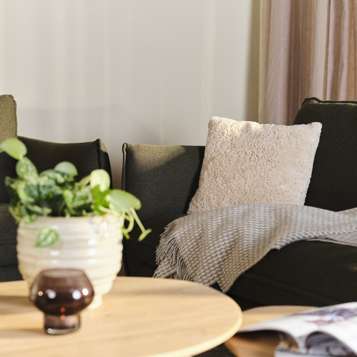 Pels putetrekk i creme i imitert pels på en mørk grønn sofa med grått pledd og plante