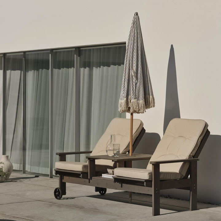 Brafab Iduna hagepute i beige på en solseng i mørkt tre med stripet parasoll på en terrasse