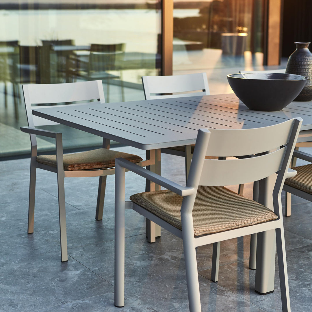 Brafab Frisk sittepute i gråbrun på hvite aluminiumsstoler ved spisebord på terrasse med glassvegg