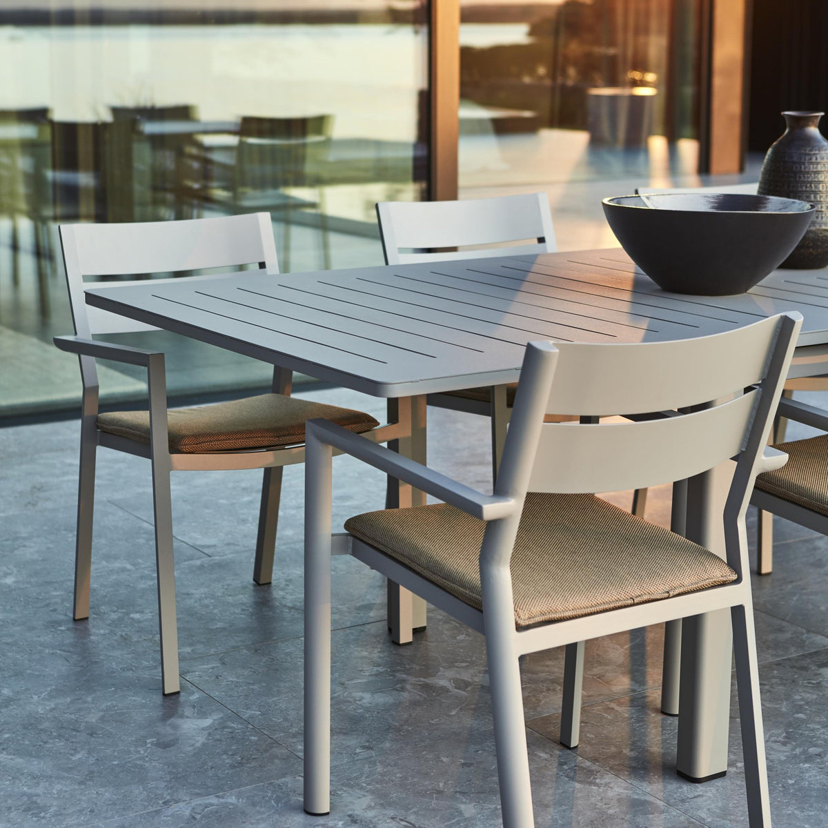 Brafab Frisk sittepute i gråbrun på hvite aluminiumsstoler ved spisebord på terrasse med glassvegg