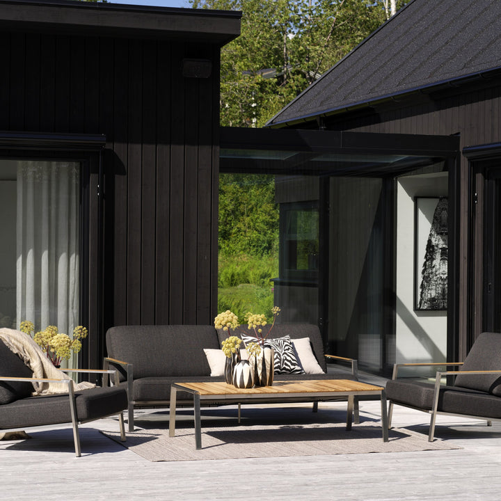 Brafab Naos utesofa i børstet stål med sorte puter og teakbord på terrasse ved sort hus
