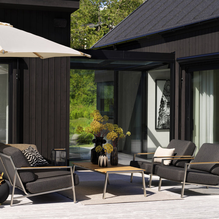 Brafab Naos utesofa med antrasittgrå puter på terrasse ved mørkt hus, med teakbord og parasoll