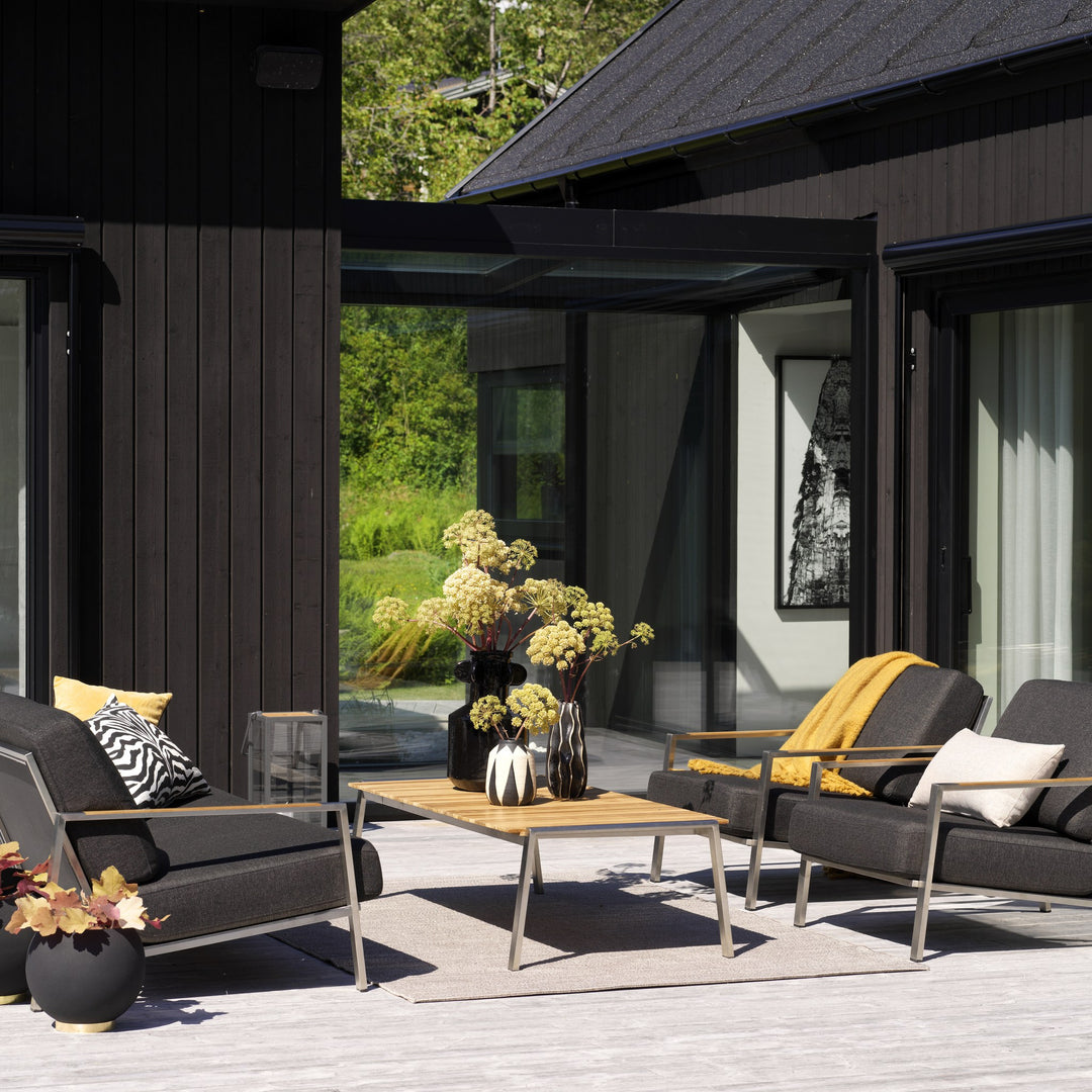 Brafab Naos utesofa med antrasittgrå puter og gult pledd på solrik terrasse med teakbord