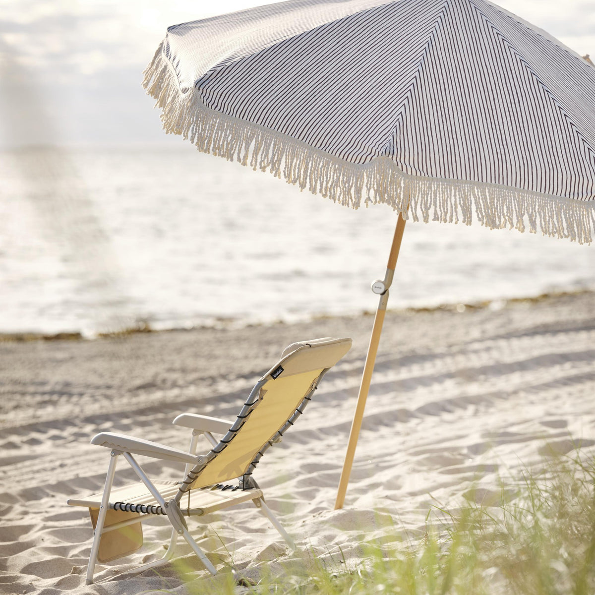 Brafab Gatsby parasoll i blå og hvite striper med frynser på stranden, gul strandstol