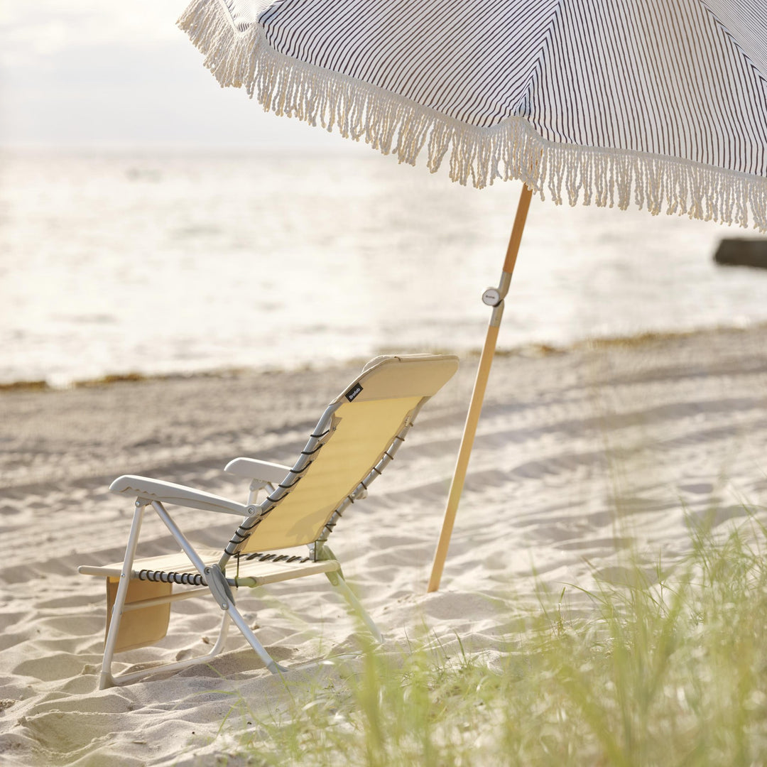 Brafab Gatsby parasoll med blå striper og frynser på sandstrand, gul strandstol og hav bak