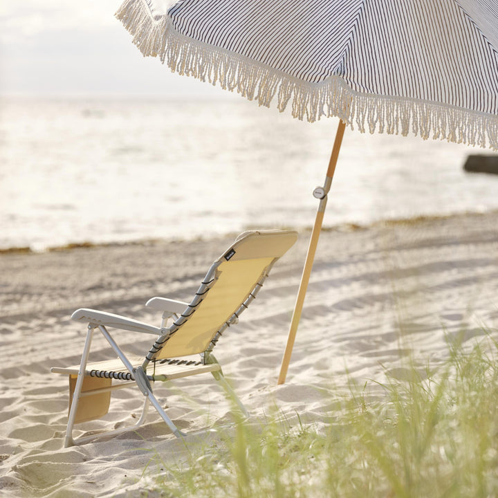 Brafab Gatsby parasoll med blå striper og frynser på sandstrand, gul strandstol og hav bak