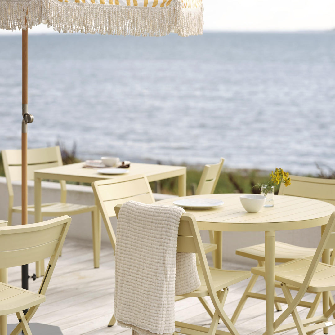 Brafab Gatsby parasoll med frynser over lysegult utemøblement på terrasse ved sjøen