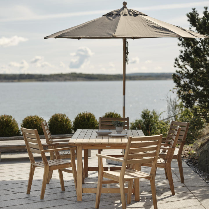 Brafab Paliano parasoll i gråbeige over teakbord med stoler på terrasse ved sjøen