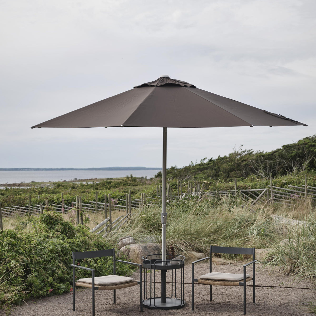 Brafab Abriola parasoll i gråbrun, åpnet over to stoler og rundt bord ved strand med strandgress