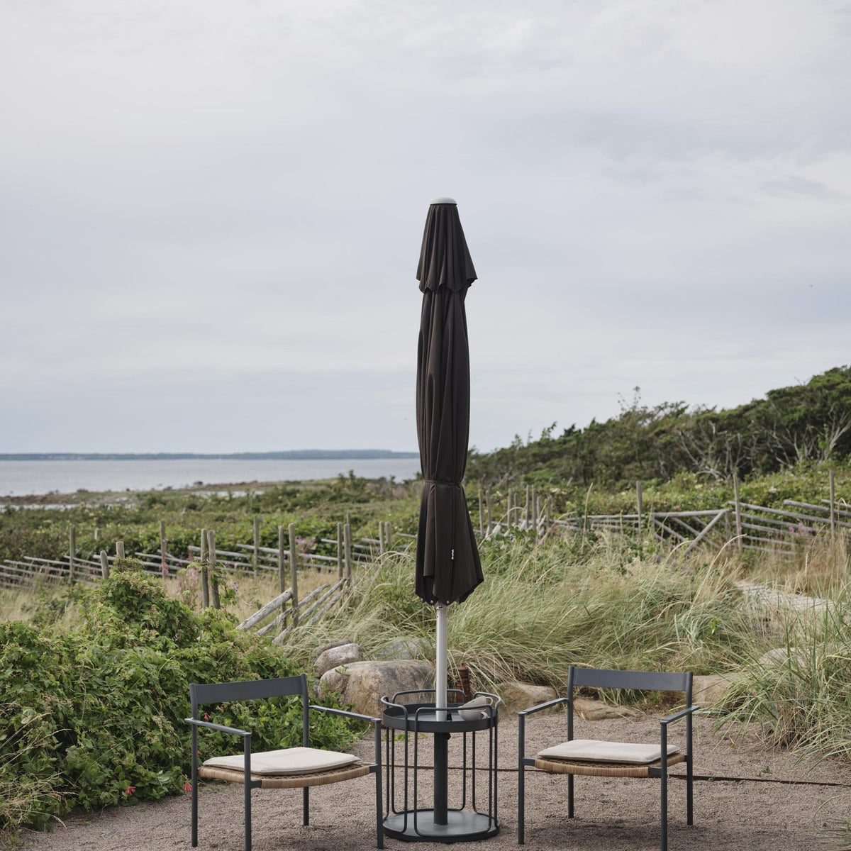 Brafab Shady parasollfot i antrasitt med sammenslått parasoll, to stoler ved strand med havutsikt