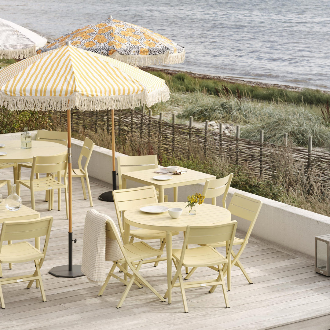 Brafab Wilkie klappstol i lysegul rundt bord på terrasse ved sjøen med stripete parasoller