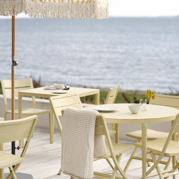 Brafab Wilkie klappstoler i lys gul på terrasse ved sjøen, med hvitt pledd og dekket bord