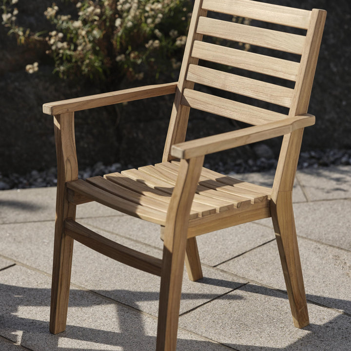Brafab Keros spisestol i teak med armlener på steinheller, grønn plante i bakgrunnen
