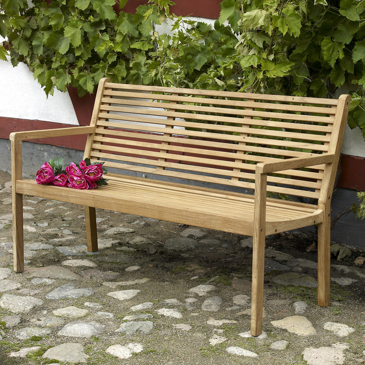 Brafab Canberra hagebenk i teak med rosa pioner på setet, foran murvegg med vinranker og brostein