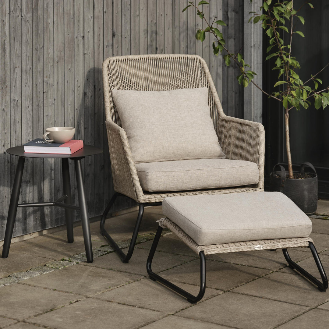 Brafab Midway loungestol i beige tau med pute og fotskammel på steinhelle ved tregjerde