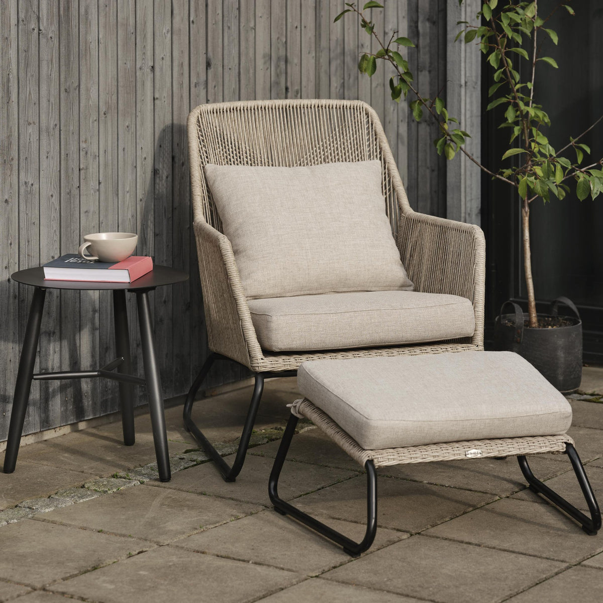 Brafab Midway fotskammel i beige på terrasse foran loungestol, sidebord med kopp, bøker og plante