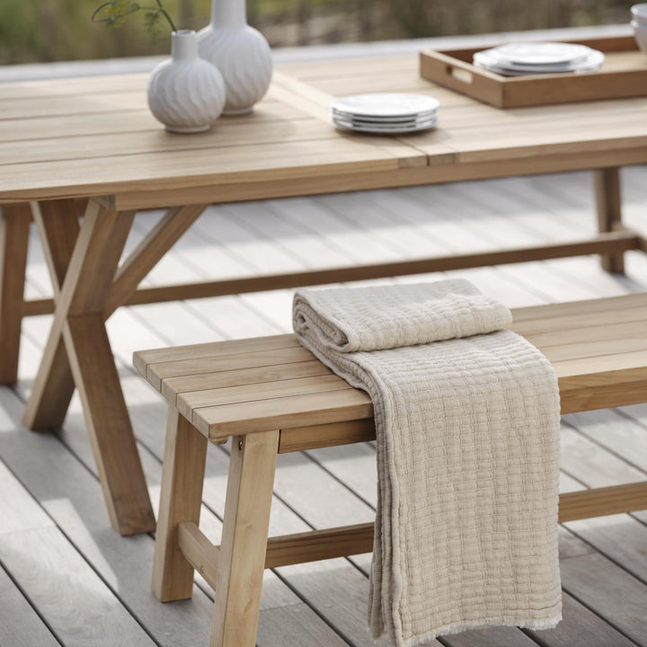 Brafab Keros benk i teak med beige pledd, ved teakbord på tredekk med vaser og tallerkener