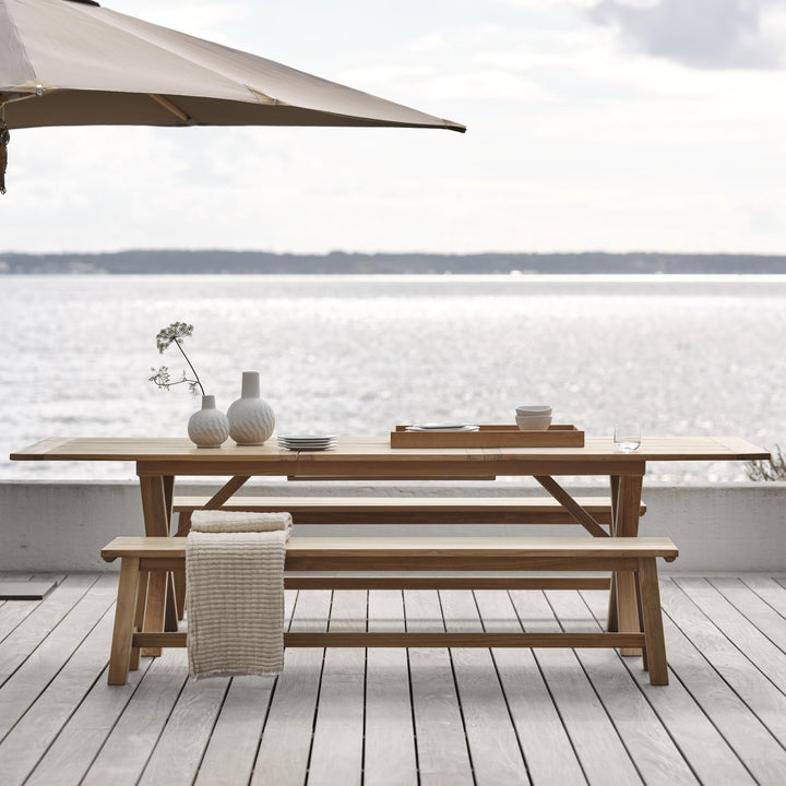 Brafab Keros benk i teak ved langbord på bryggeterrasse mot sjøen, med parasoll og hvite vaser.