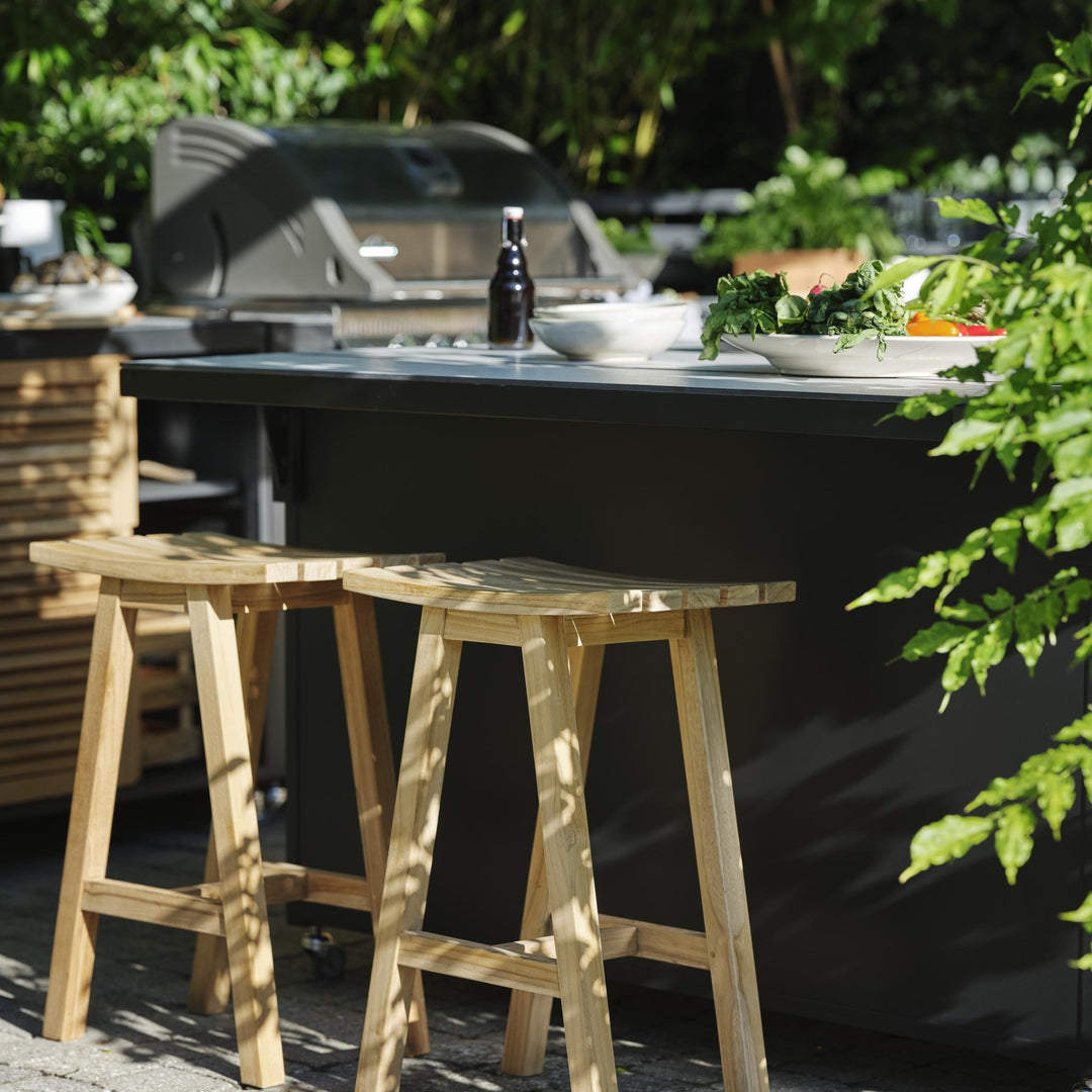 Brafab Keros barstol i teak ved utekjøkken med grill, omgitt av grønne planter.