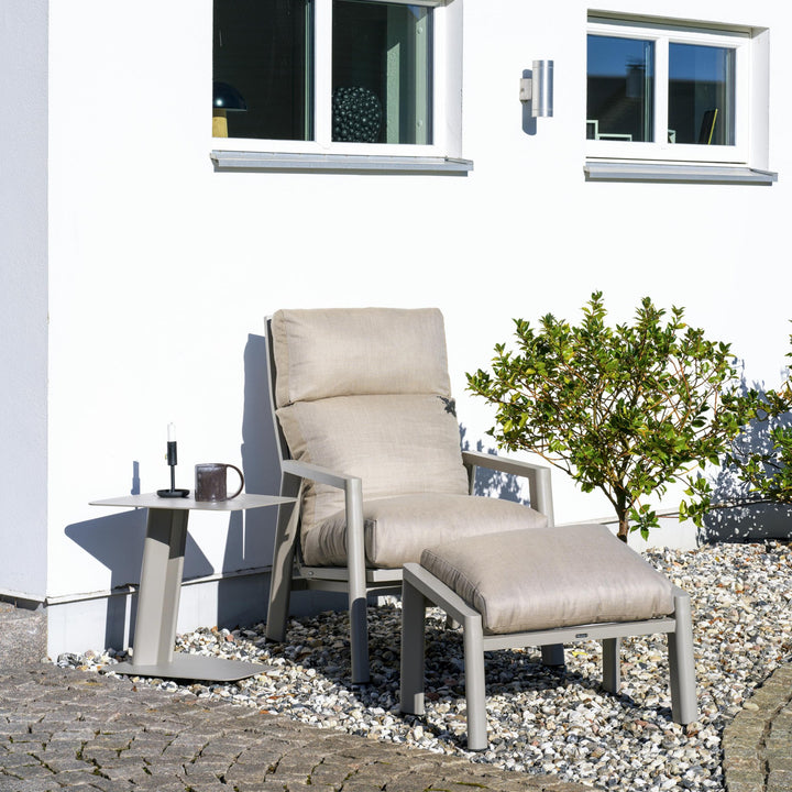 Brafab Slide hagestol i khaki med beige puter og fotskammel, ved hvit husvegg på steinbed.