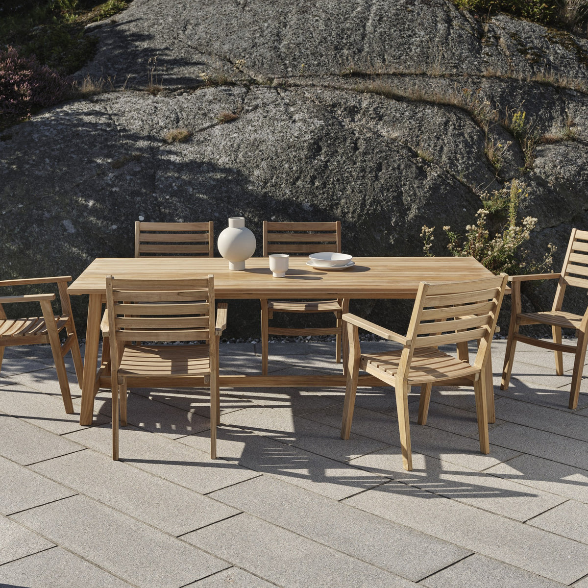 Brafab Keros spisebord i teak med stoler på steinheller foran fjellvegg, hvit vase og kopper