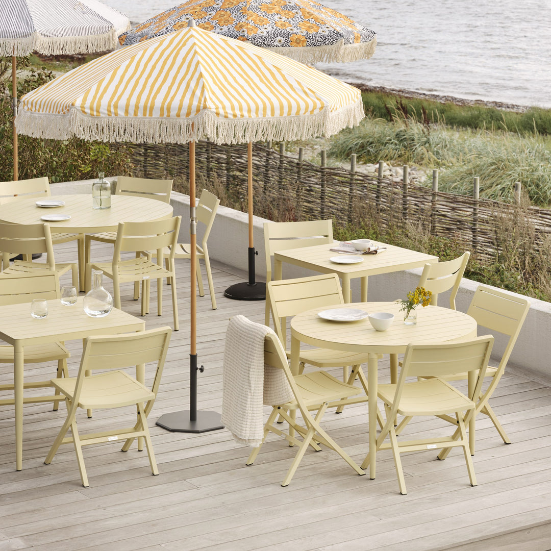 Brafab Bigby spisebord i lys gul med stoler på terrasse med stripet parasoll ved sjøen