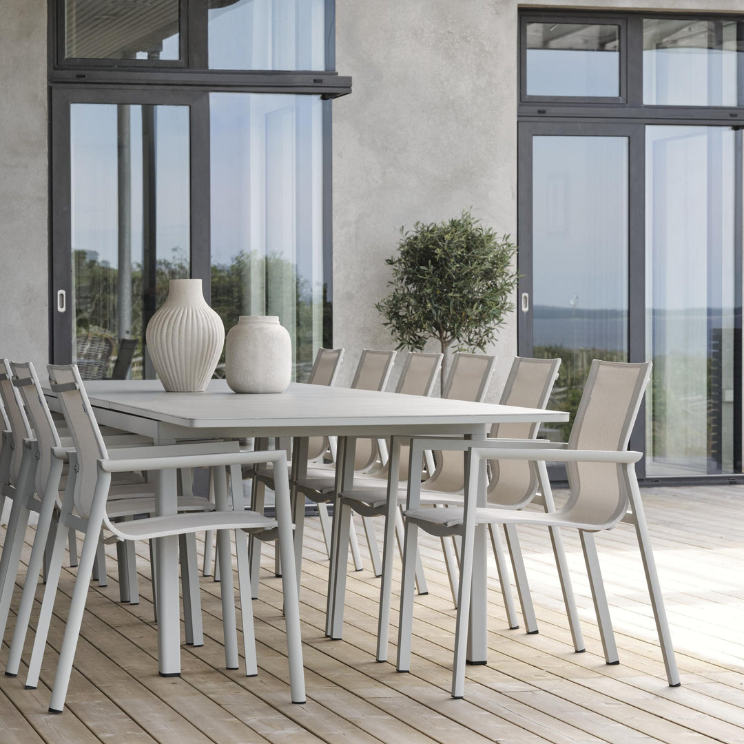 Brafab Lomma XL spisebord i lys grå med beige tekstilstoler på terrasse, vaser og oliventre, sjøutsikt