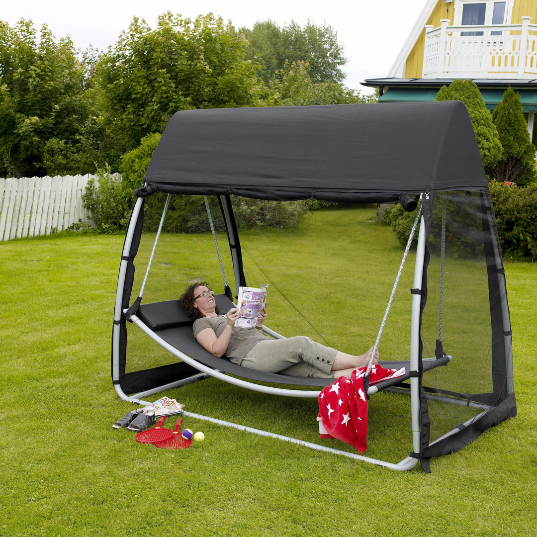 Brafab Silas hammock i sort med tak og myggnett på gressplen, person ligger og leser, hage rundt