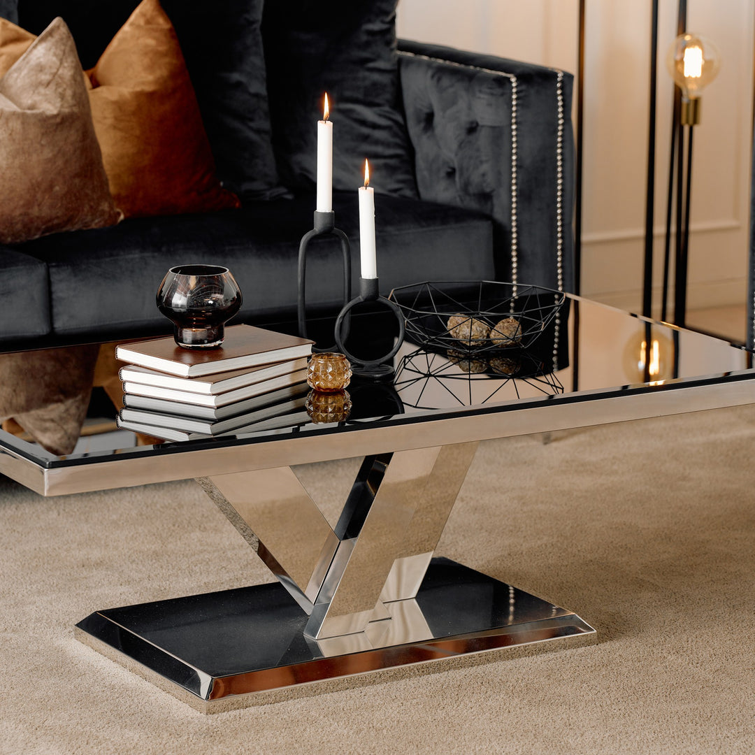 Messina sofabord med sort glassplate og blank V-formet krombase ved sort sofa