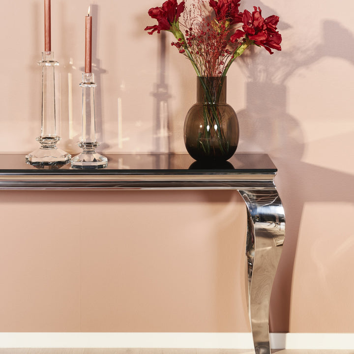 Lavezzi konsollbord med sort glassplate og svungent krombein mot rosa vegg, med lysestaker og blomster