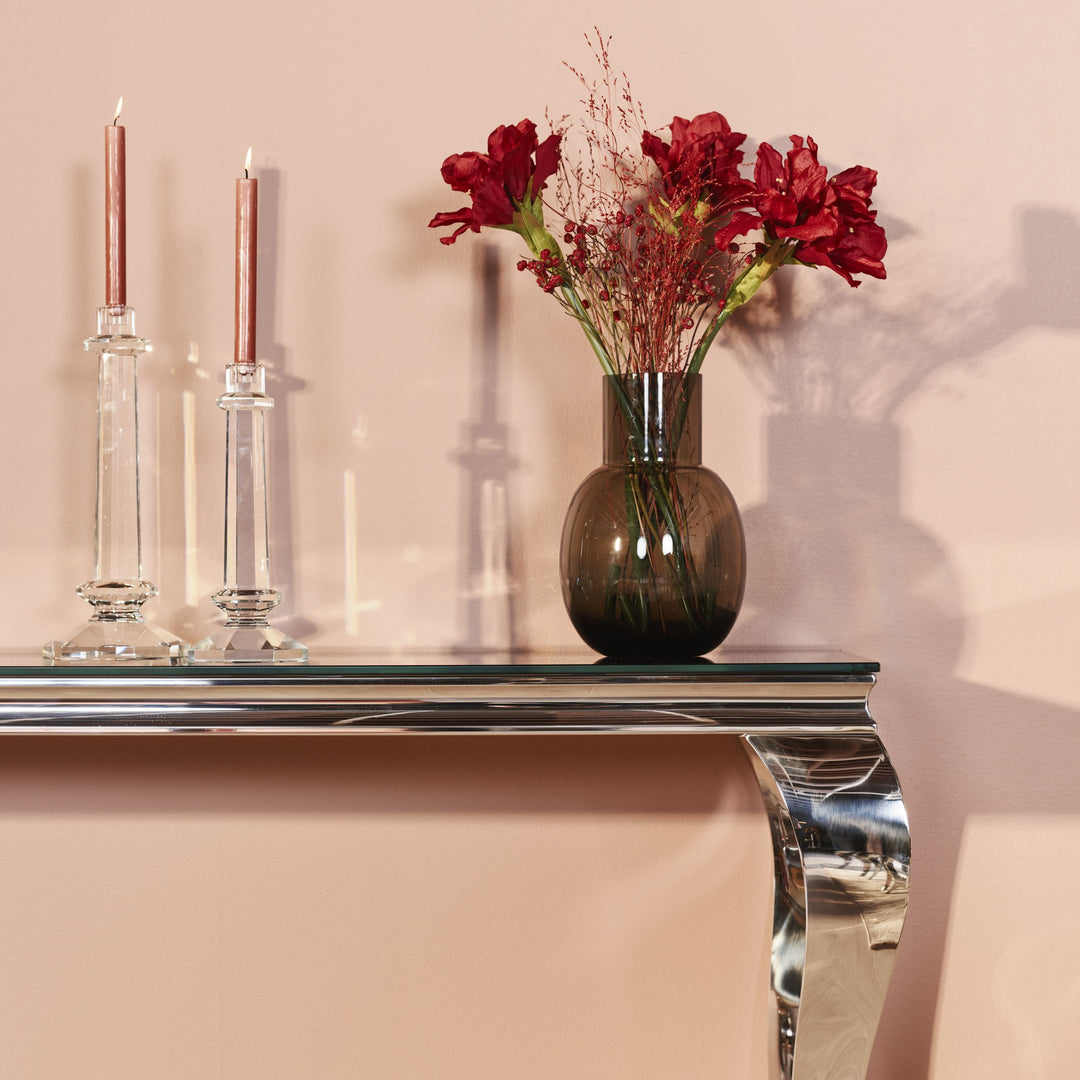 Lavezzi konsollbord i sort glass med forkrommet ben mot rosa vegg, med lysestaker og røde blomster