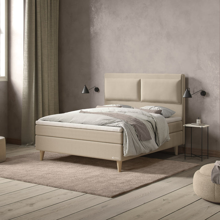 Svane Zophy kontinentalseng i beige med eikeben mot taupe vegg med vegglamper og puff