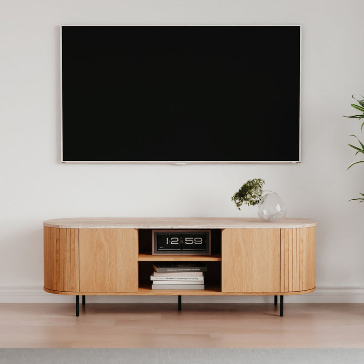 Fabia tv-benk i eik med riflet front og travertinplate, under veggmontert TV med flipklokke og grønn plante