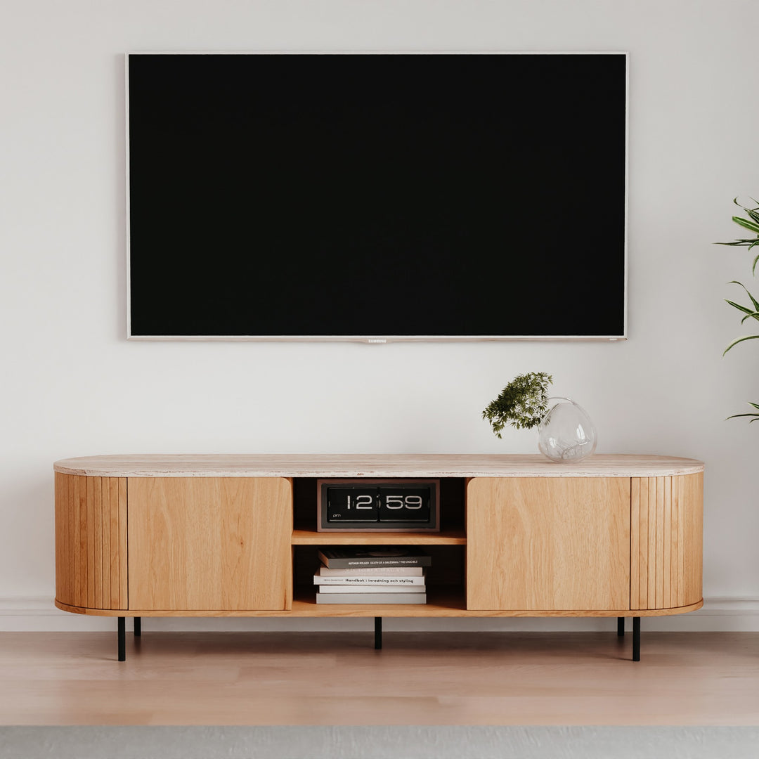Fabia tv-benk i eik med riflede, avrundede sider og travertinplate, under veggmontert TV i lyst stuemiljø