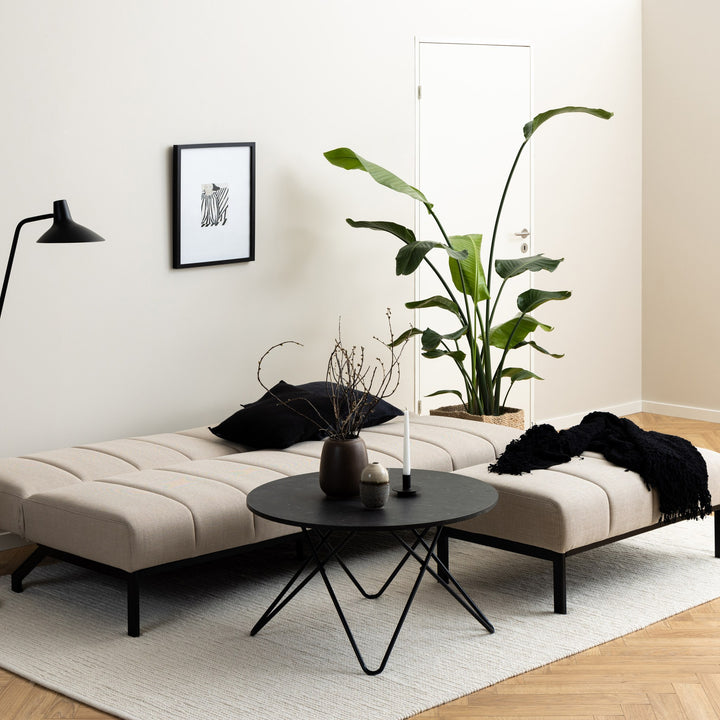Caix sovesofa i beige stoff, utslått i lys stue med svart sofabord, grønn plante og teppe