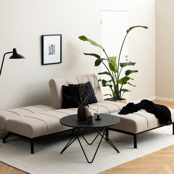 Caix sovesofa i beige stoff i minimalistisk stue med rundt svart sofabord og stor grønn plante