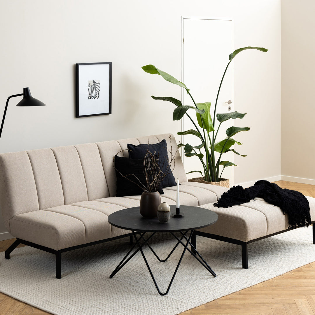 Caix sovesofa i beige stoff i lys stue med stor grønnplante, sort gulvlampe og rundt sofabord