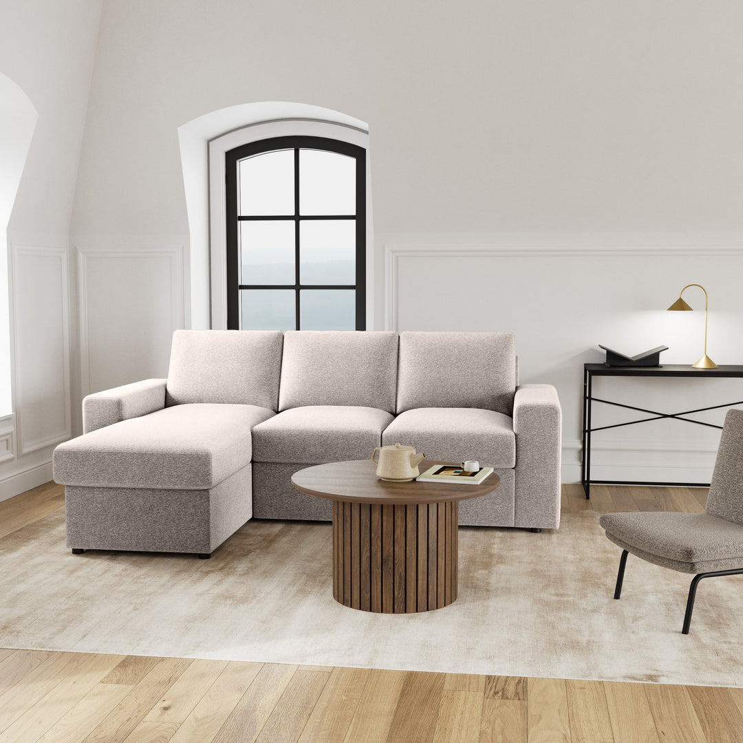 Conan sovesofa i lys beige med sjeselong, rundt trebord og buevindu i lyst rom