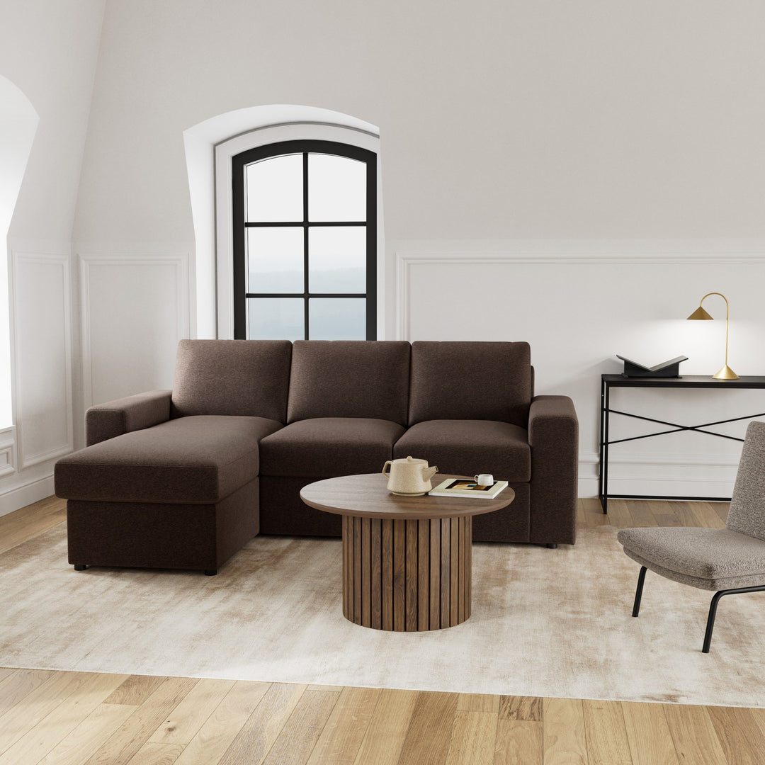 Conan sovesofa i mørk brun med sjeselong, rundt trebord og buevindu i lyst rom