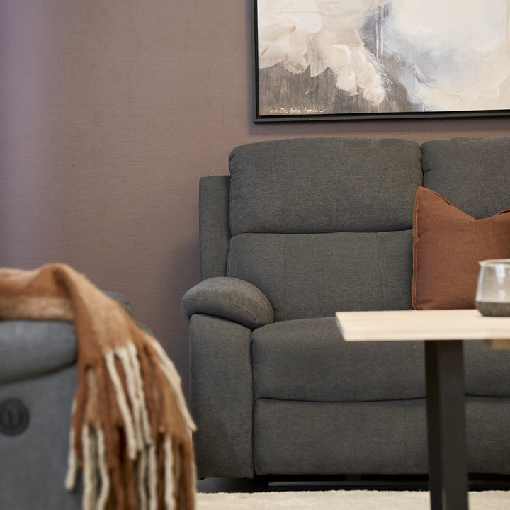 Mora reclinersofa i mørk grå med rustfarget pute, mot lilla vegg.