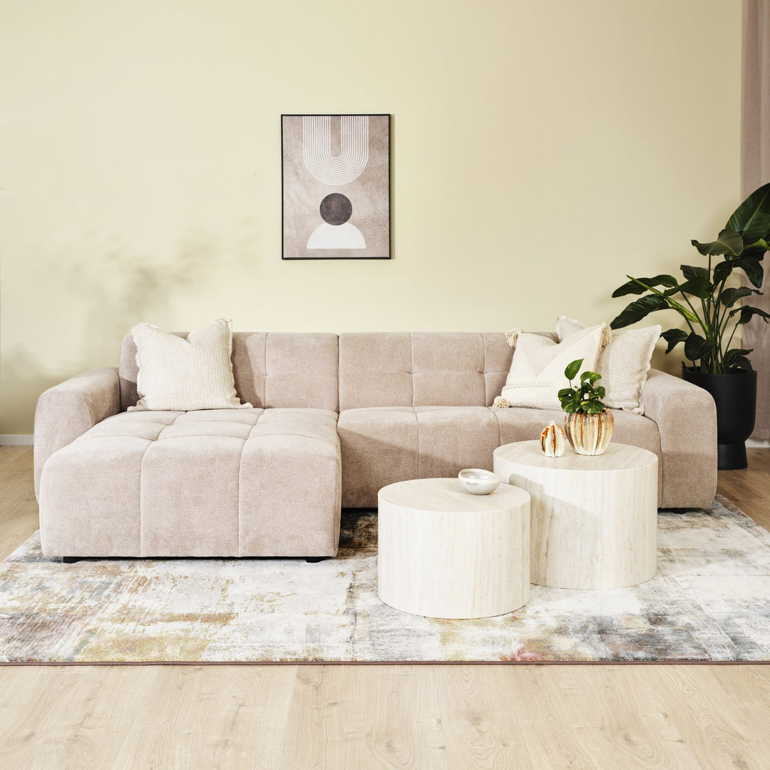 Columbia hjørnesofa i sandfarget stoff med sjeselong i stuemiljø med rundbord, plante og kunst