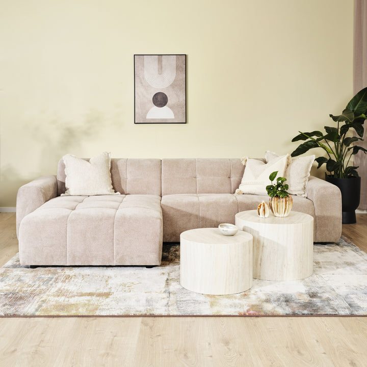 Columbia hjørnesofa i sandfarget stoff med sjeselong i stuemiljø med rundbord, plante og kunst