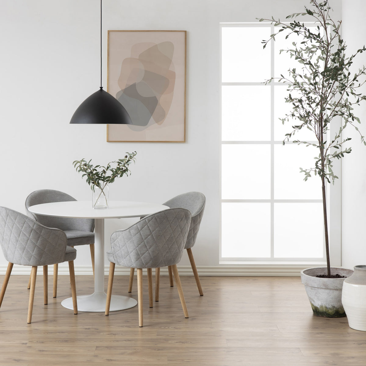 Ibiza spisebord i hvitt med rund plate og søylefot, grå quiltede stoler, sort taklampe og oliventre.