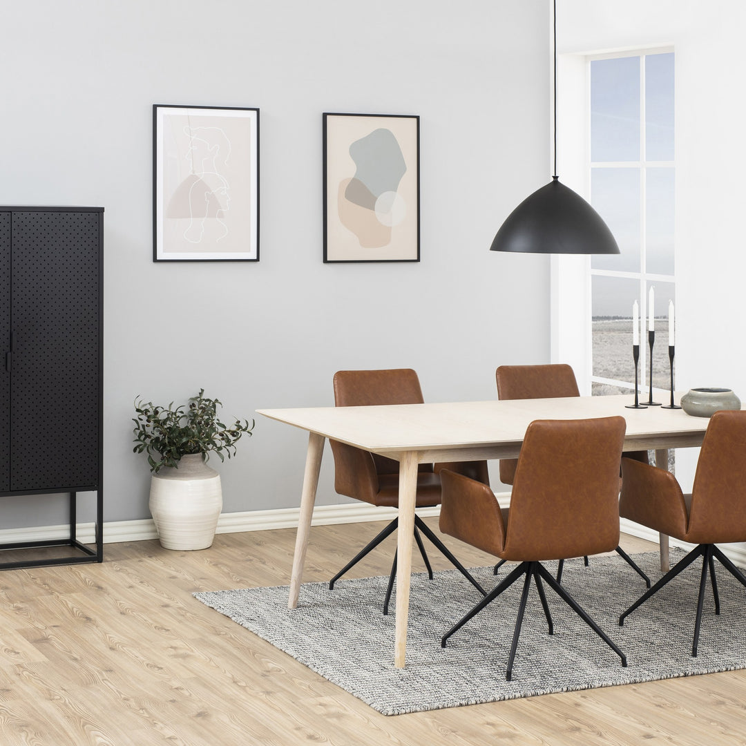 Naya spisestol i cognac kunstskinn med svarte dreieben, ved lyst spisebord med plante og pendel