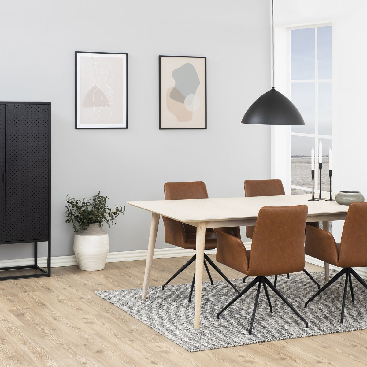 Naya spisestol i cognac kunstskinn med svarte dreieben, ved lyst spisebord med plante og pendel