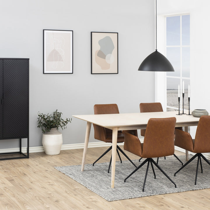Naya spisestol i cognac kunstskinn med svarte dreieben, ved lyst spisebord med plante og pendel