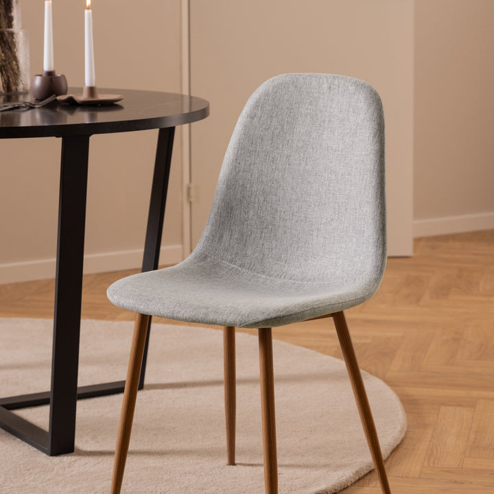 Celia spisestol i lys grå stoff med eikeben, ved sort rundt spisebord med stearinlys på beige teppe