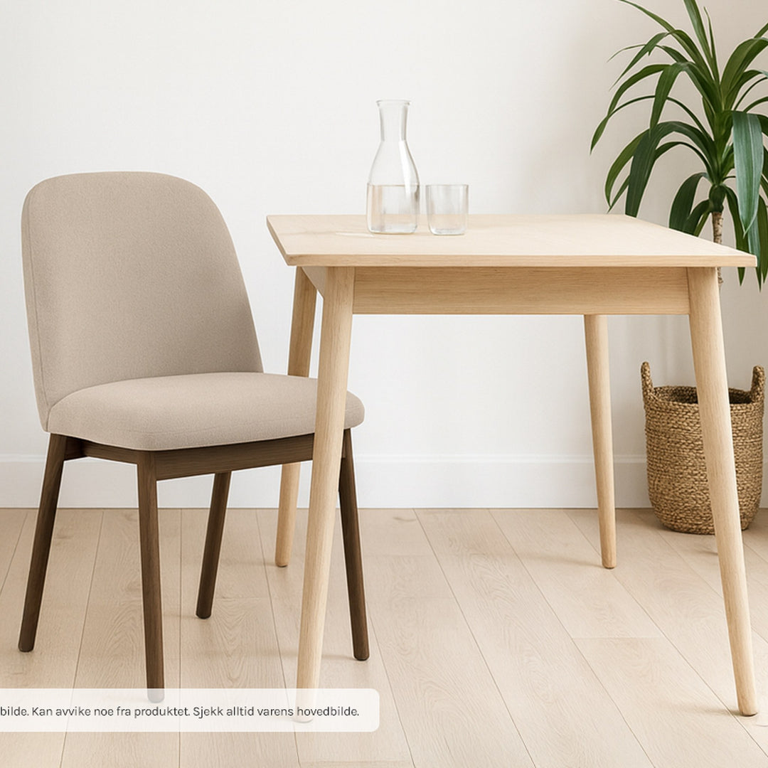 Leo spisestol i beige stoff med mørke trebein, ved spisebord i lys eik med plante og kurv.